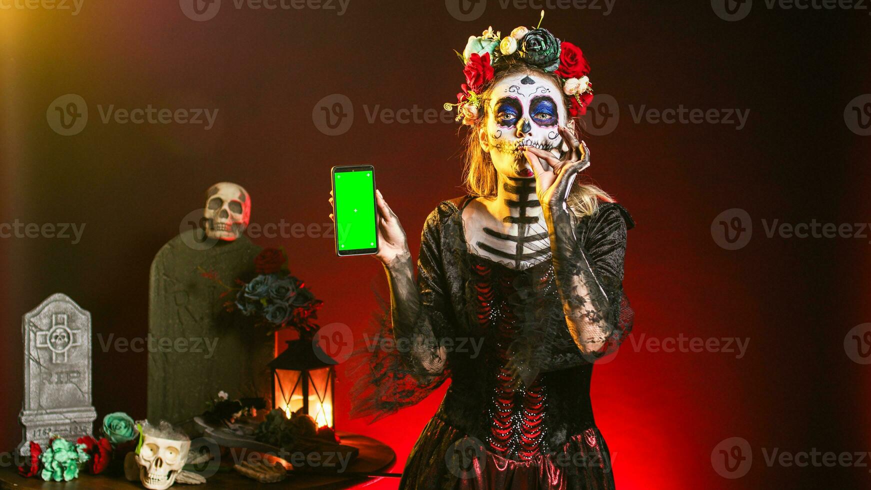 Creepy female model holding greenscreen on smartphone, using mobile phone with isolated mockup template. Woman dressed as santa muerte showing blank chroma key copyspace display. photo