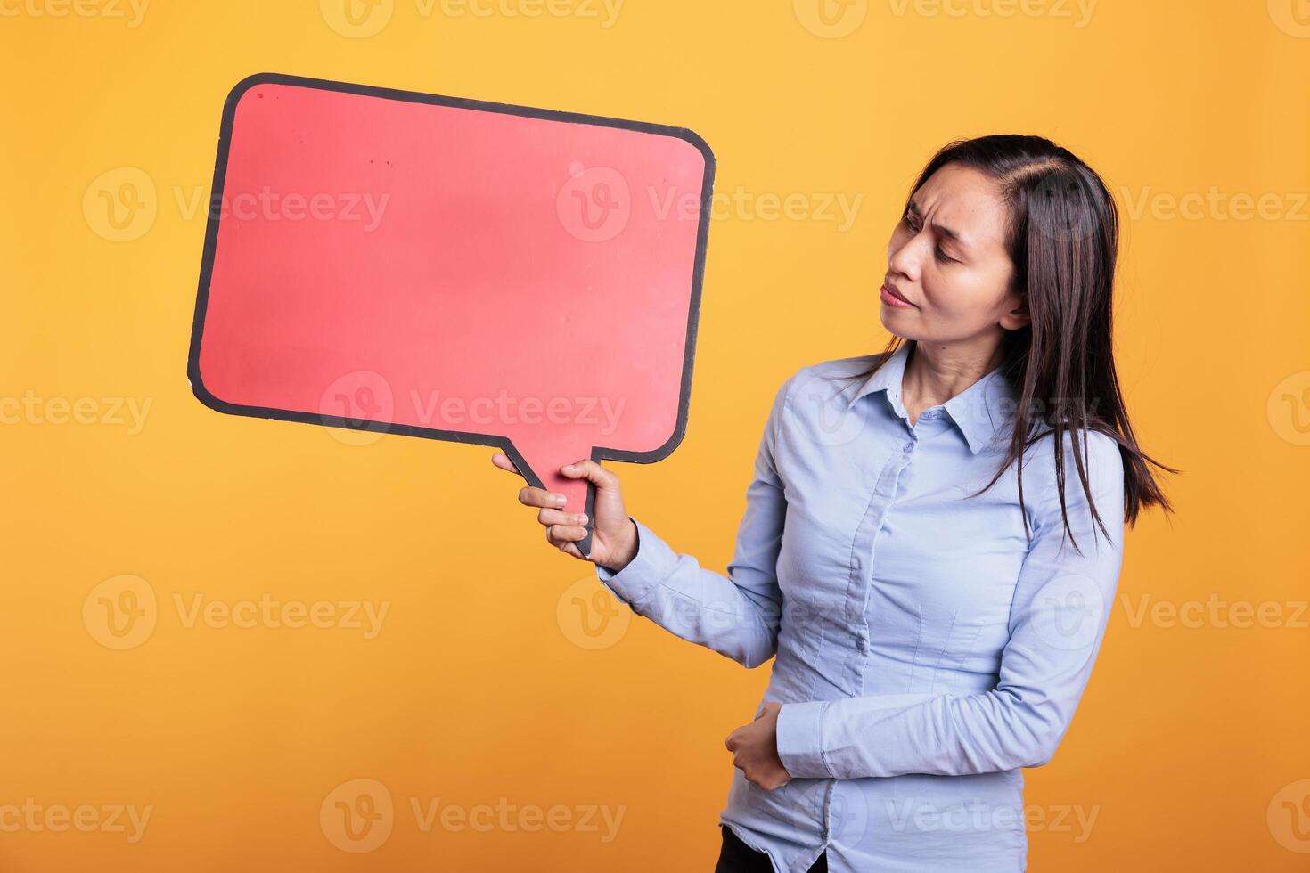 Filipino woman holding empty red speech bubble with copy space, advertising product. Cheerful young adult standing in studio over yellow background, looking at blank dialog frame with place for text photo