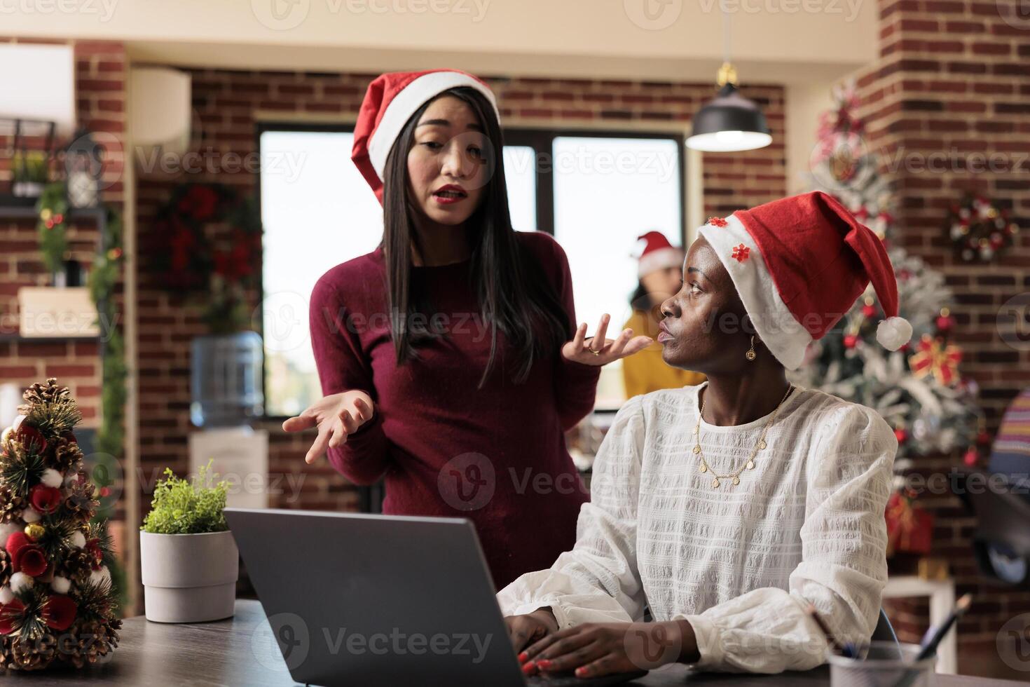 Women coworkers in santa hats discussing and brainstorming on laptop in decorated workspace. Asian and african american colleagues doing teamwork in winter festive coworking space photo