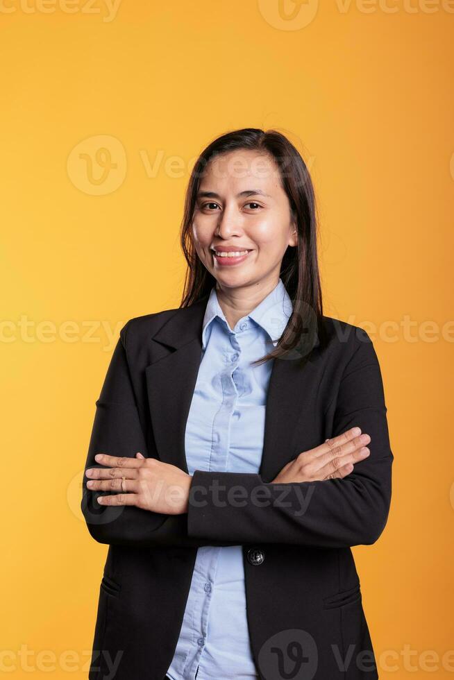 Portrait of cheerful asian woman in formal suit posing in studio over yellow background, holding arm crossed during photoshot. Smiling carefree model with joyful expression looking at camera photo