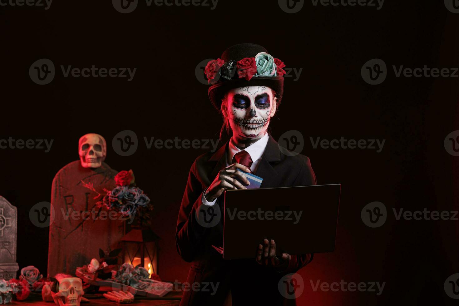 Young adult doing online shopping with skull make up, using credit card on laptop to make payment. Wearing santa muerte lady of death costume to celebrate mexican halloween holiday. photo