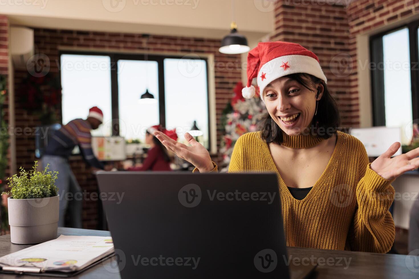 Smiling woman employee using teleconference app on laptop to discuss project deadline in christmas decorated office. Cheerful project manager in santa hat greeting coworkers in videocall photo