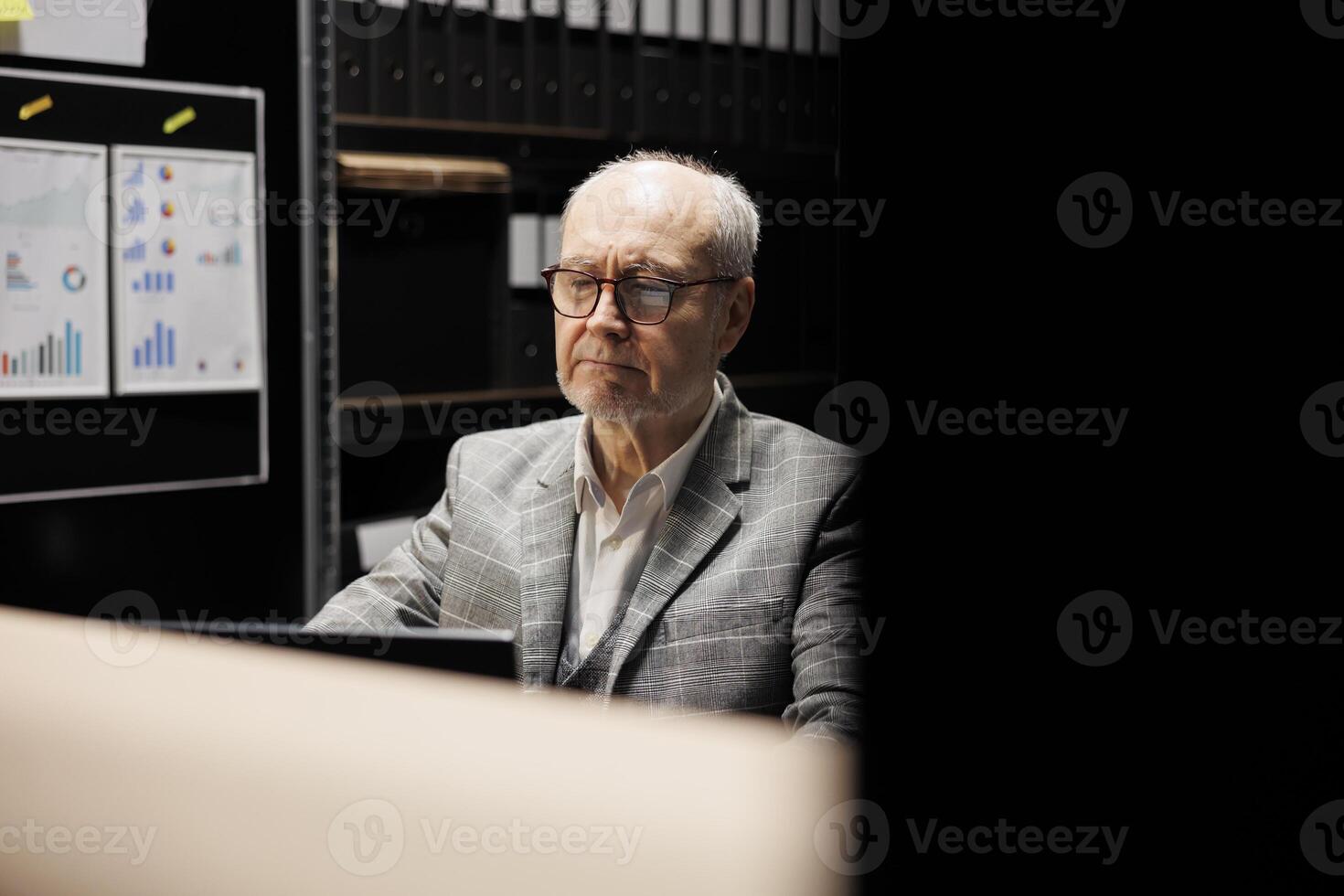 Senior businessman executive sitting at office desk, inspecting quarterly company results analysis data paperwork on laptop. Accountancy workplace filled with document folders on cabinet shelves photo