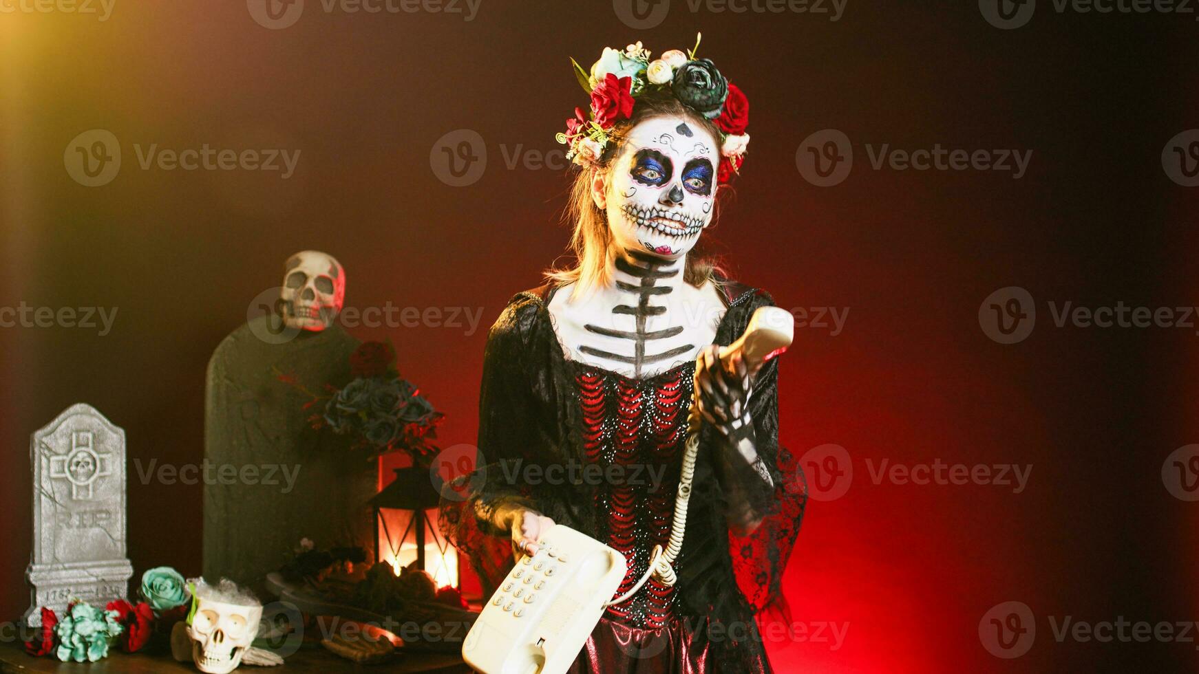 Spooky woman answering landline phone with cord, talking on office telephone while she has skull make up and body art. Portraying santa muerte lady of death on dios de los muertos celebration. photo