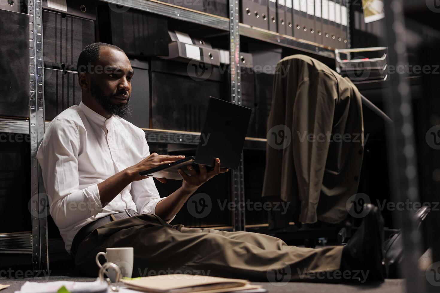 Private detective conducting investigation using laptop while sitting on floor surrounded by police report papers. Detective analyzing crime case information on computer in evening photo