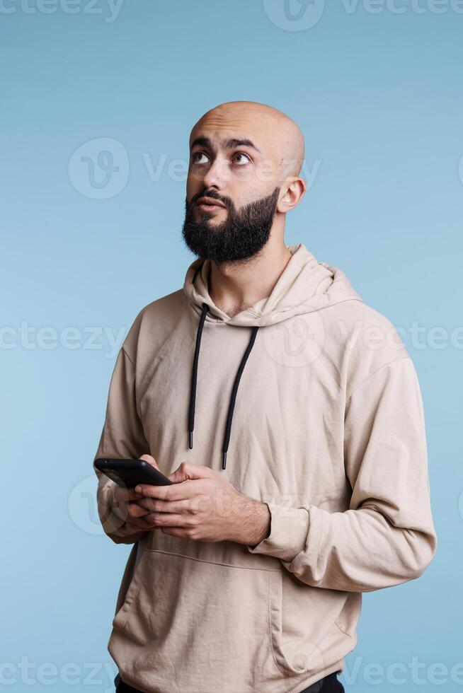 pensativo árabe hombre mensajes de texto en teléfono inteligente y utilizando social medios de comunicación red con pensativo expresión. joven persona en duda mientras pensando acerca de mensaje antes de enviando en móvil teléfono foto