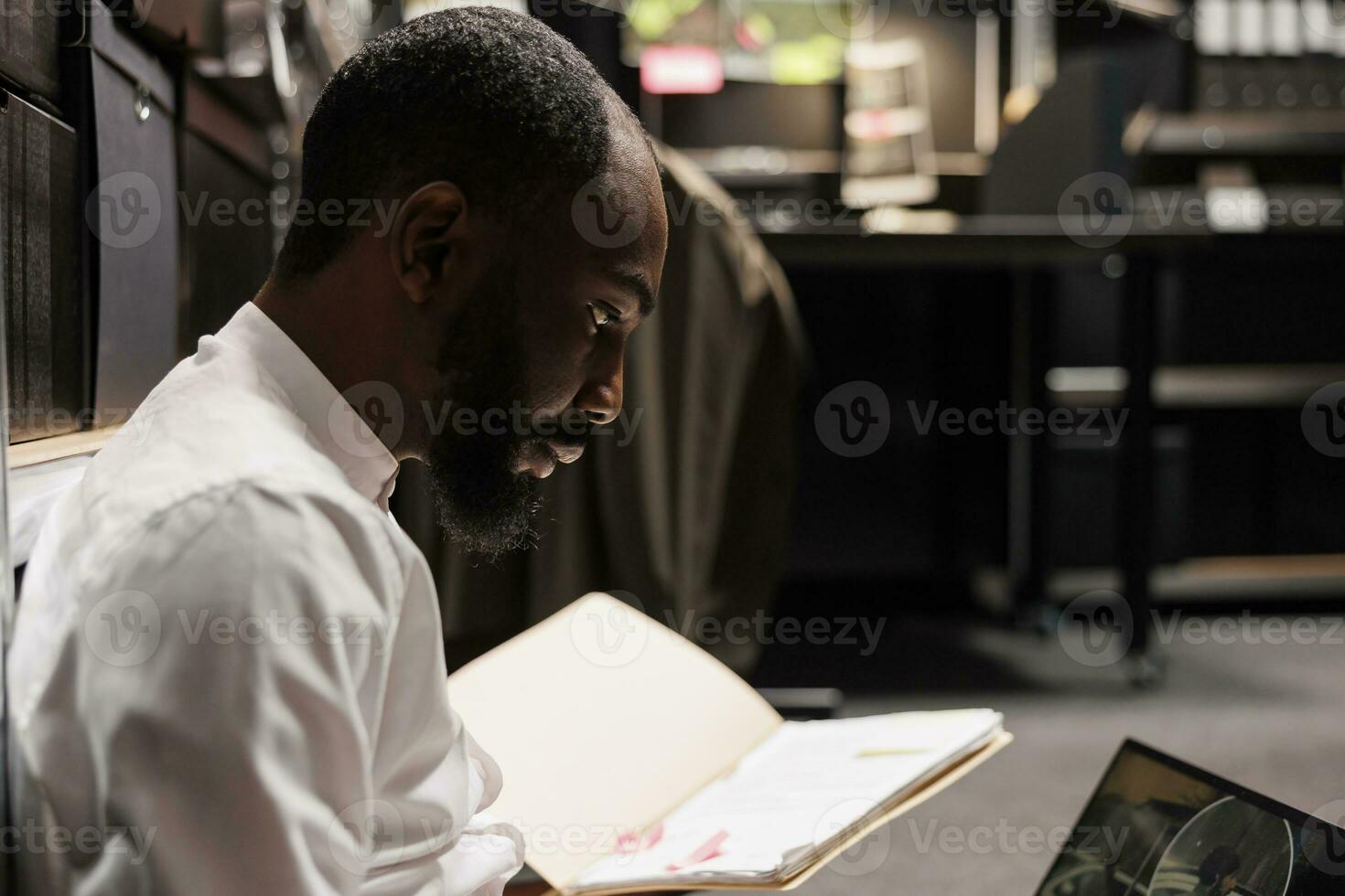 Policeman reading crime case file and analyzing photos on laptop. African american concentrated detective conducting investigation, studying forensic report and crime scene photographs