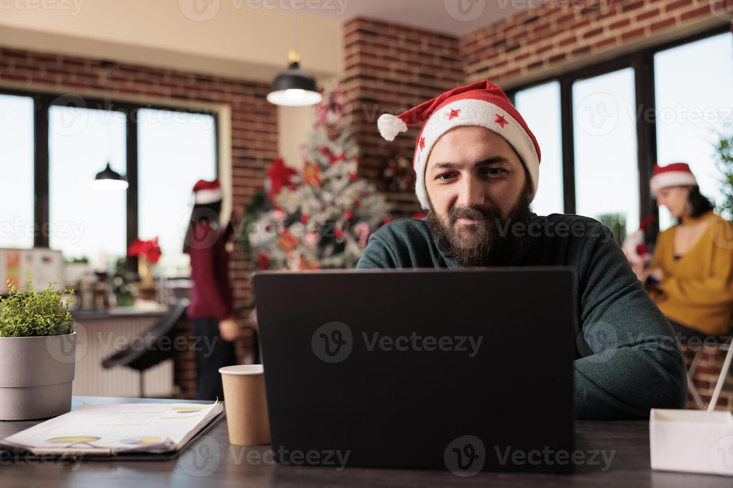 Smiling company employee checking startup project report on laptop while working in office at christmas eve. Caucasian man in santa hat using computer at festive decorated workplace photo