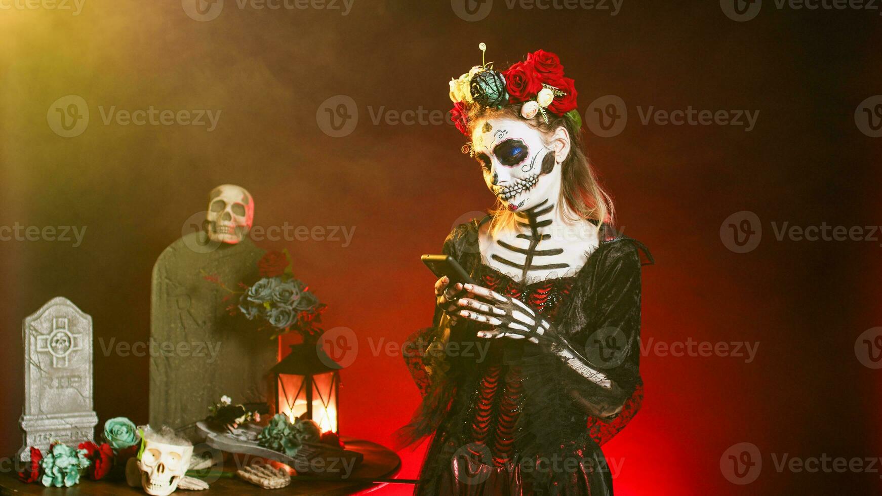 Glamorous scary woman using smartphone to browse internet website over black background. Santa muerte model portraying horror goddess of death on mexican holiday celebration in studio. photo