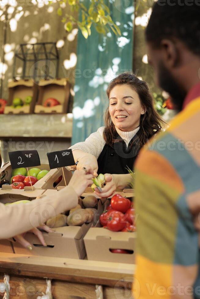 Smiling friendly organic fruits and vegetables shop owner offering customer to taste different varieties of apples at food market. Positive female farmer selling fresh local homegrown products photo