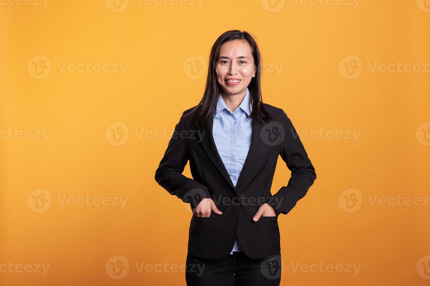 Positive woman standing with hands in pocket posing over yellow background, enjoying studio shot. Smiling confident model having joyful expression, smiling at camera during work break photo