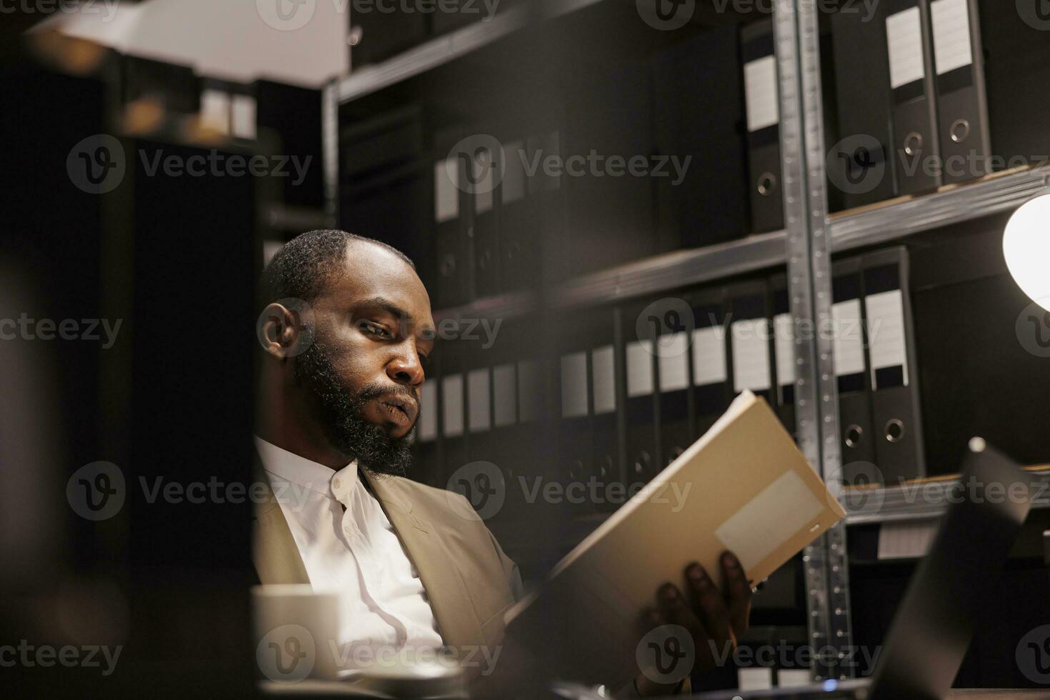 Exhausted african american detective solving complex crime case and reading folder with police reports. Tierd investigator working late, studying archival records information photo