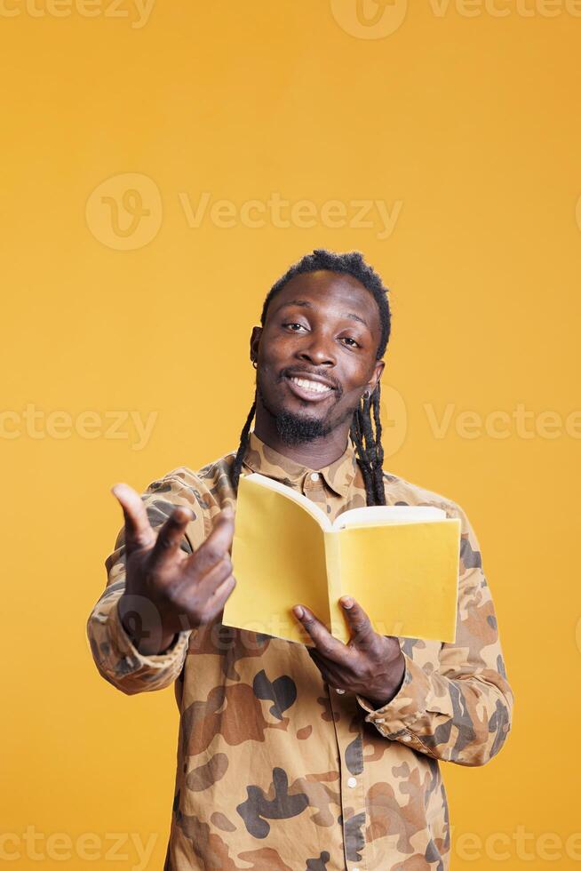 Smiling african american man enjoying fiction storytale, holding literature book while posing in studio over yellow background. Positive clever man reading story for university homework photo