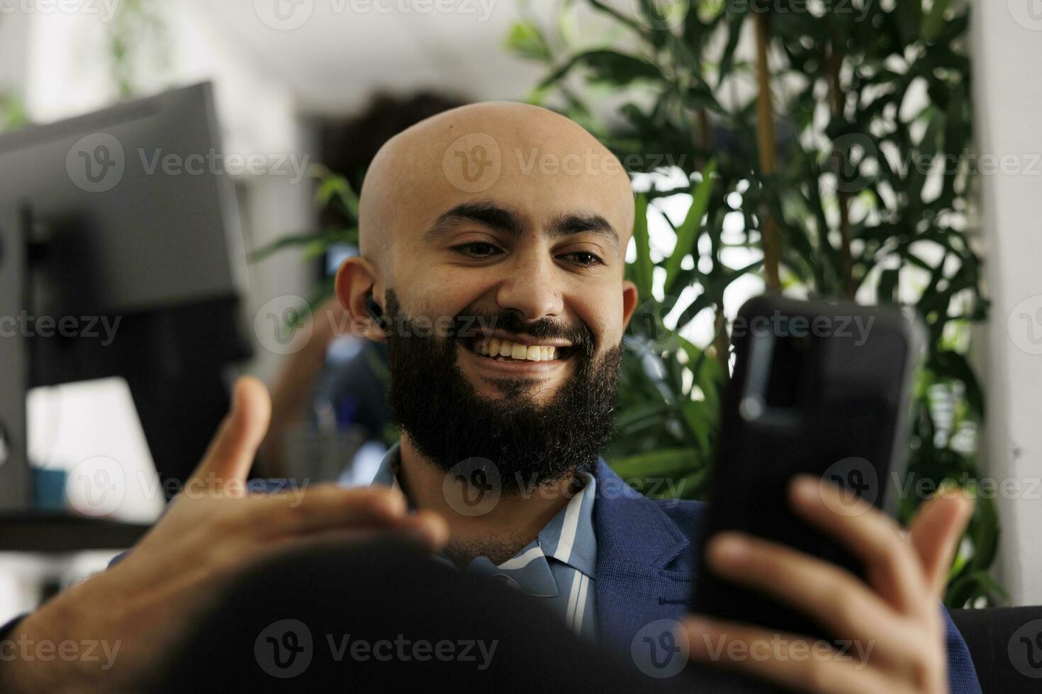 Smiling arab executive manager teleconferencing using smartphone while working in business office. Startup entrepreneur doing virtual team management on mobile phone videocall photo