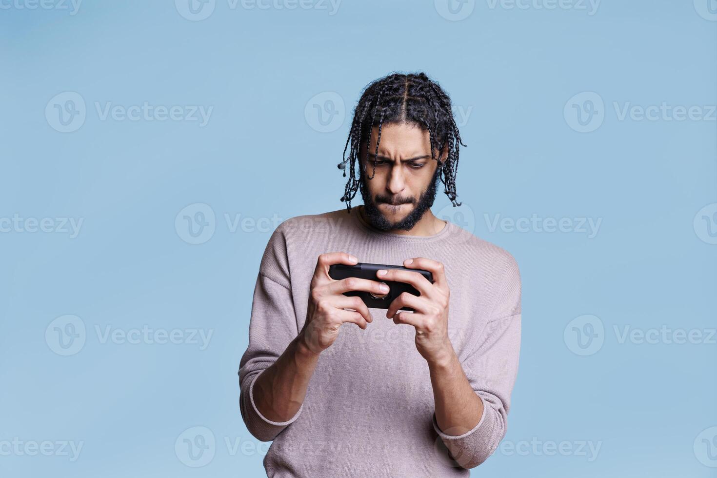 Focused arab man playing video game on smartphone with concentrated facial expression. Young gamer wearing casual clothes using mobile app for electronic leisure activity photo