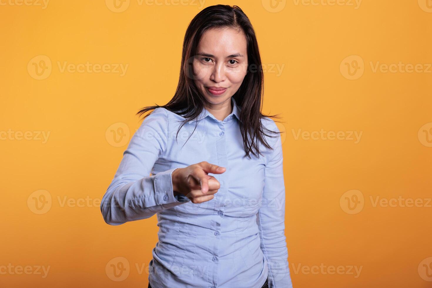 Successful asian woman pointing index finger at you, posing in studio over yellow background. Carefree smiling young adult pointing forefinger advertising promotional product. Advertisement concept photo