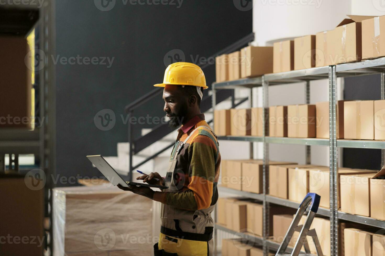 African american worker holding laptop computer, analyzing products checklist in warehouse. Storage room employee wearing protective overall while preparing customers orders before delivery goods photo