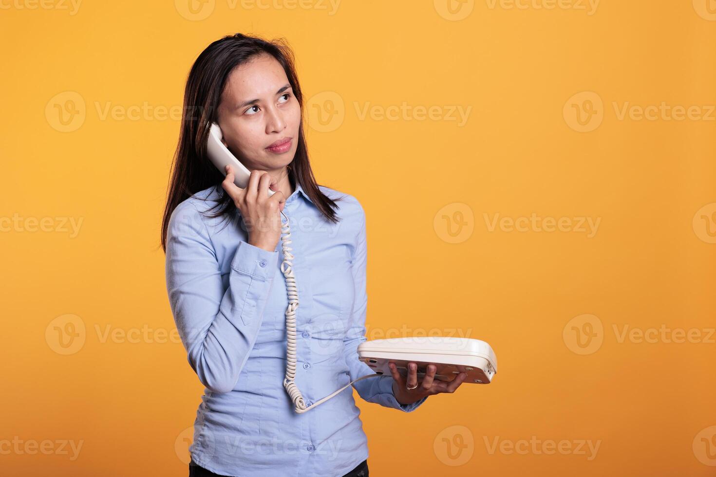Asian woman talking at landline phone with remote worker, chatting on retro telephone line with cord in studio over yellow background. Cheerful smiling young adult enjoying lifestyle discussin photo