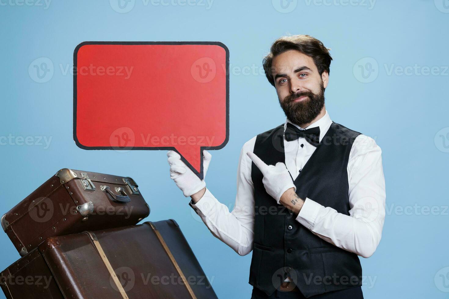 Young doorman points at speech bubble against blue background, doing marketing ad using red billboard sign with copyspace. Concierge employee holding blank isolated cardboard on camera. photo
