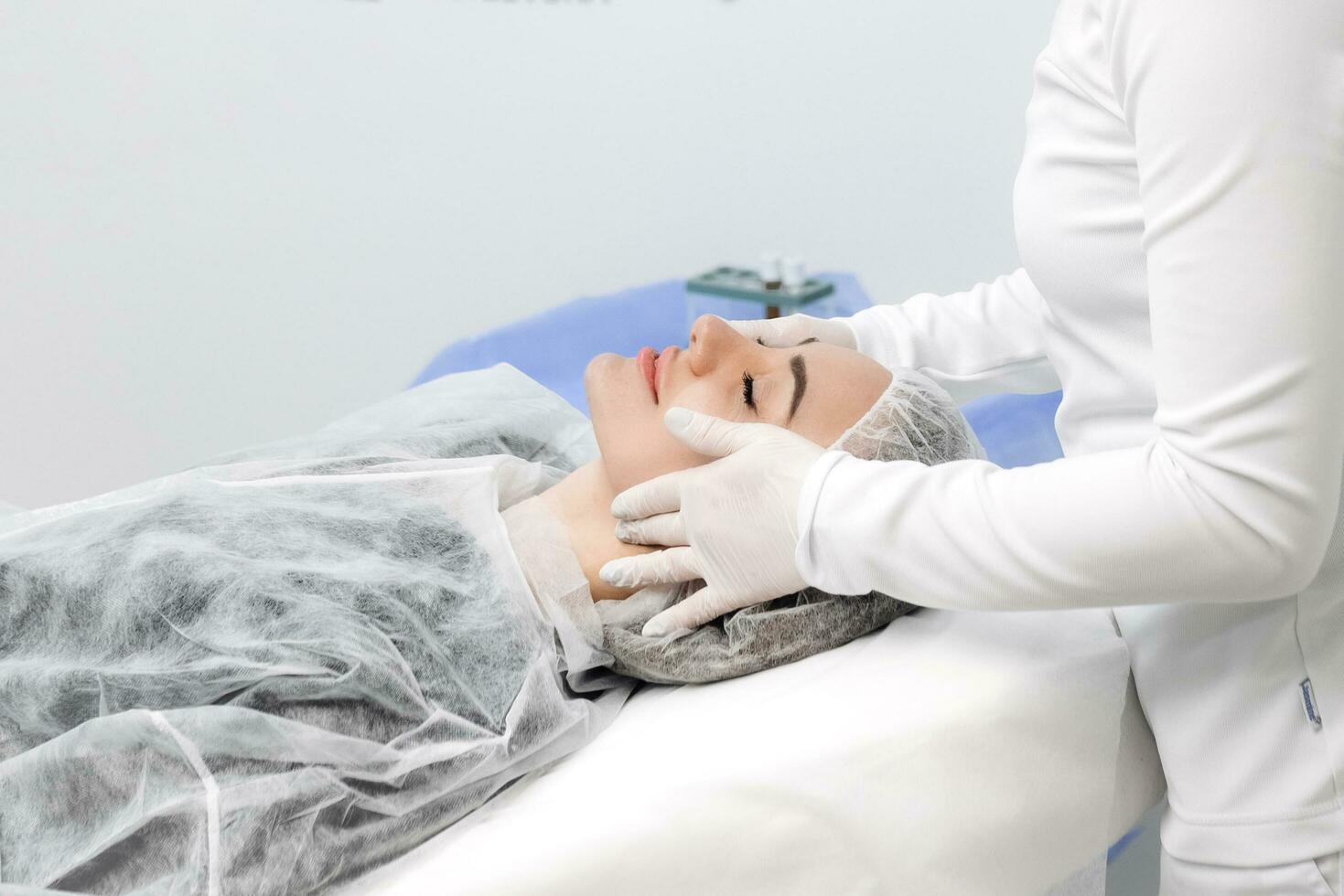 Cosmetologist is applying cream with anesthesia on patient's face skin before biorevitalization procedure. Woman in beauty clinic with doctor beautician preparing to treatment using numbing cream. photo