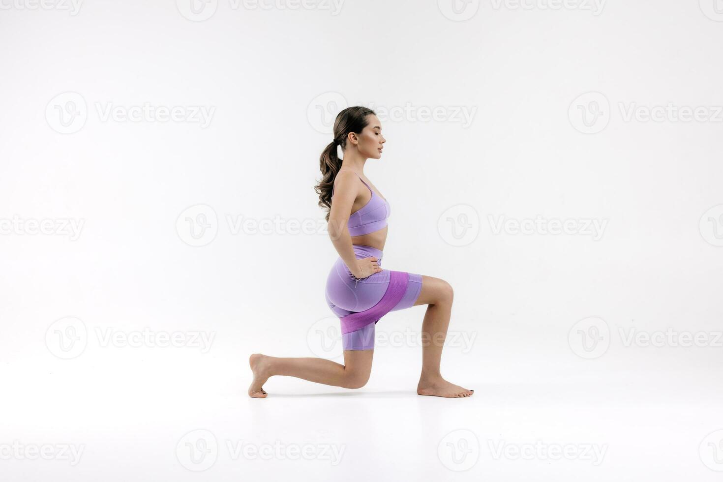 Woman doing squats in a purple sports suit with fitness elastic band on white background photo