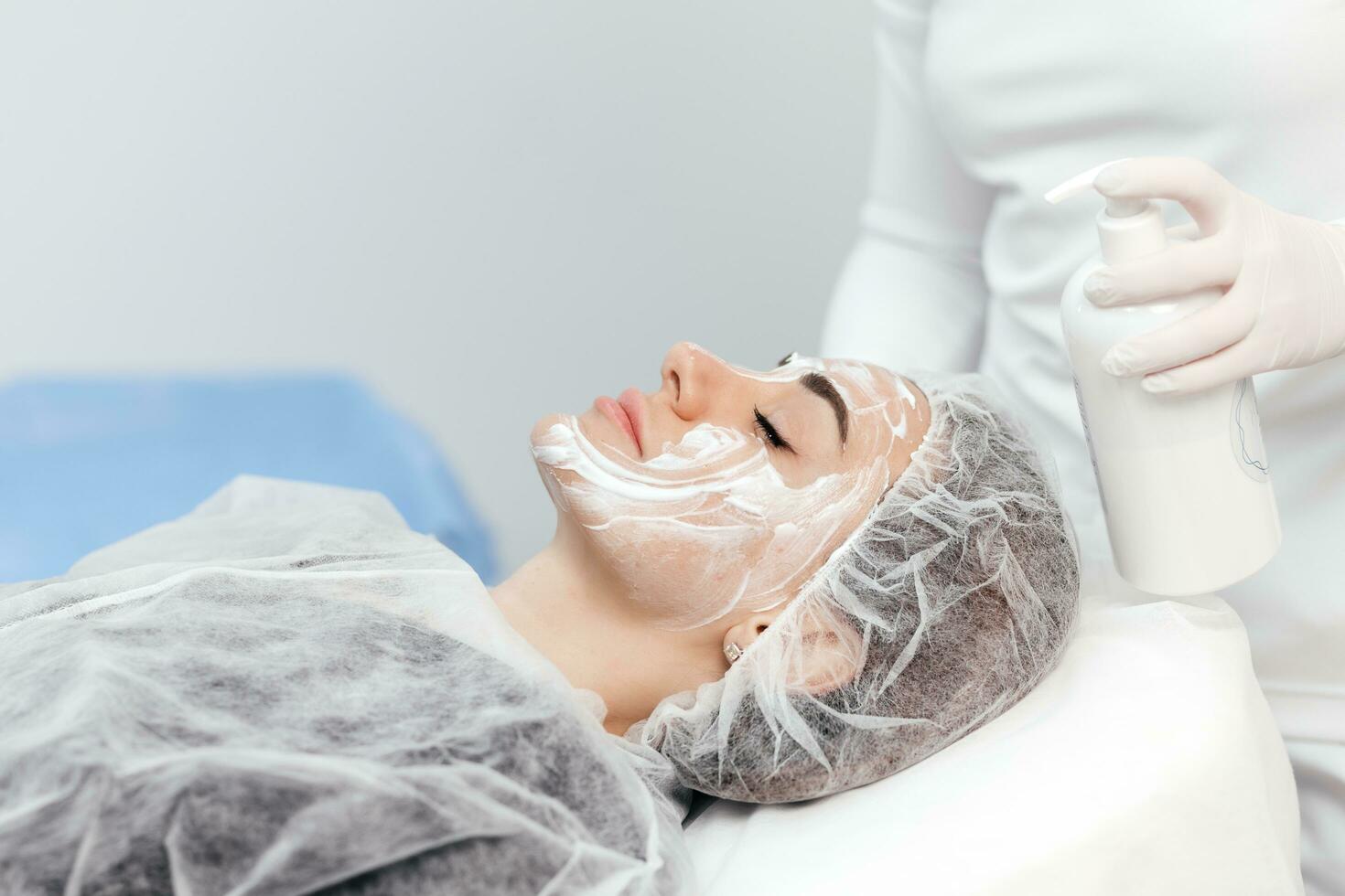 Cosmetologist is applying cream with anesthesia on patient's face skin before biorevitalization procedure. Woman in beauty clinic with doctor beautician preparing to treatment using numbing cream. photo