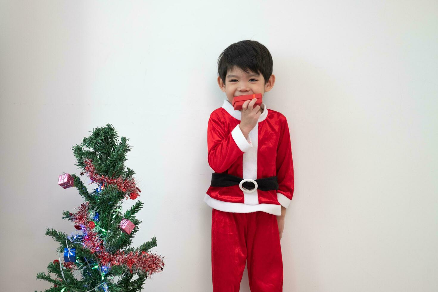Asian boy wearing Santa costume Standing and playing near A fun Christmas tree on white background. photo