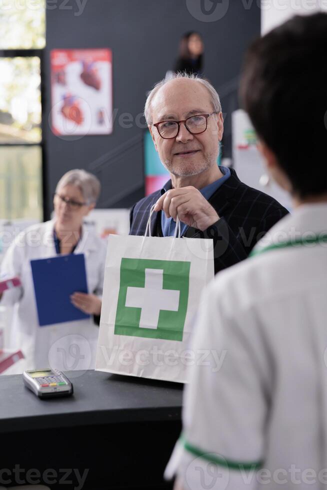 Old man taking shopping bag from pharmacy cashier at checkout, smiling to drugstore employee. Elderly customer buying prescription medicaments in medical retail store and standing at counter desk photo