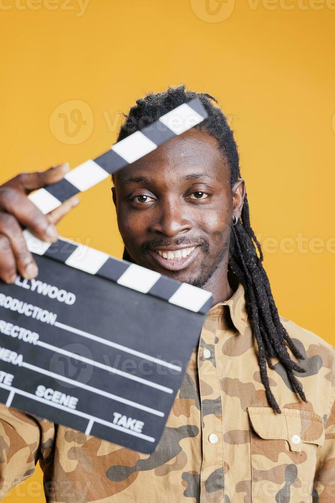 Portrait of producer holding clapperboard to cut scenes in movie making industry. African american adult presenting chalkboard to camera and smiling while having filmmaker object. Cinema concept photo