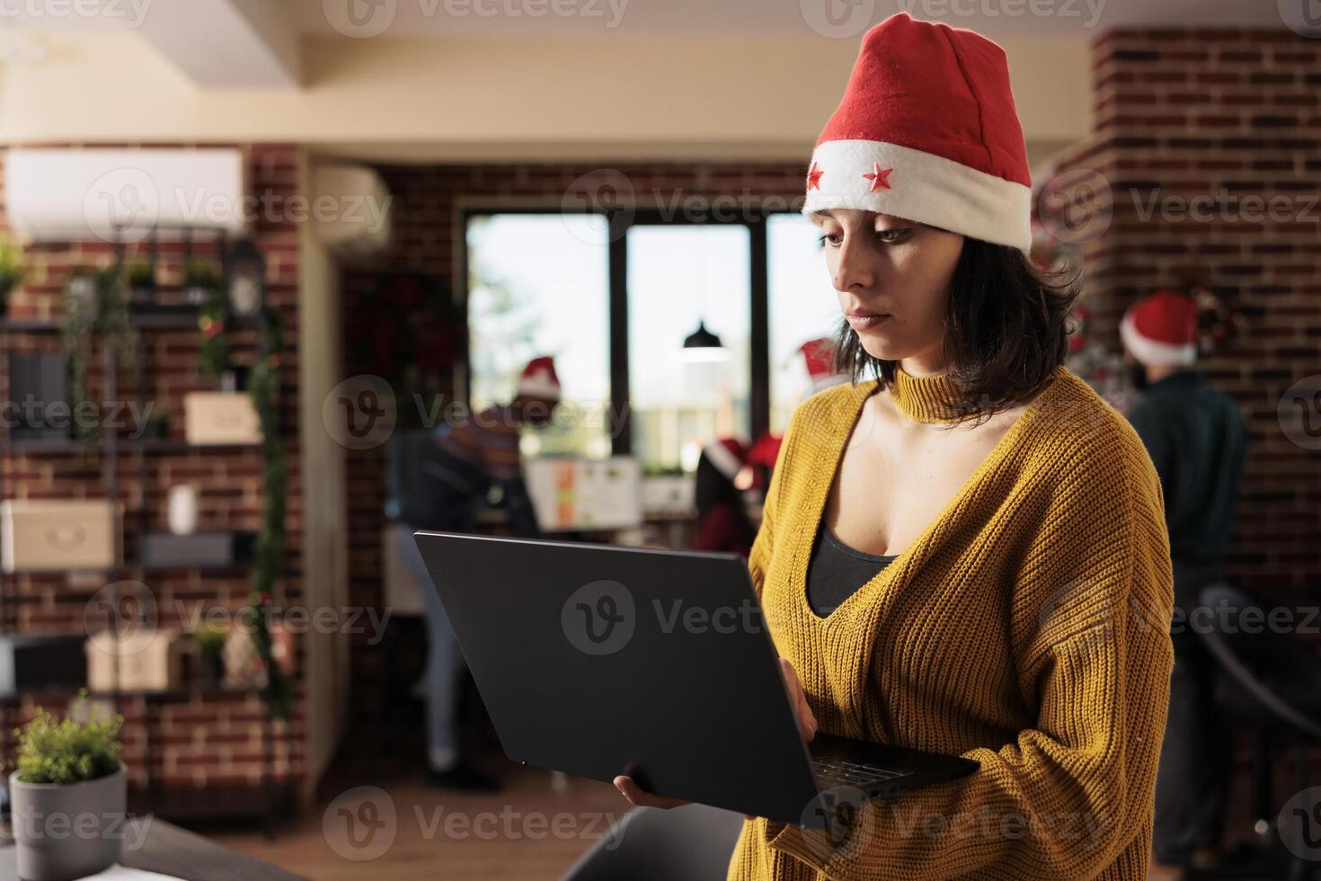 Woman project manager wearing santa hat managing tasks on laptop while standing in winter festive office. Employee holding portable computer and working for deadline at christmas eve photo