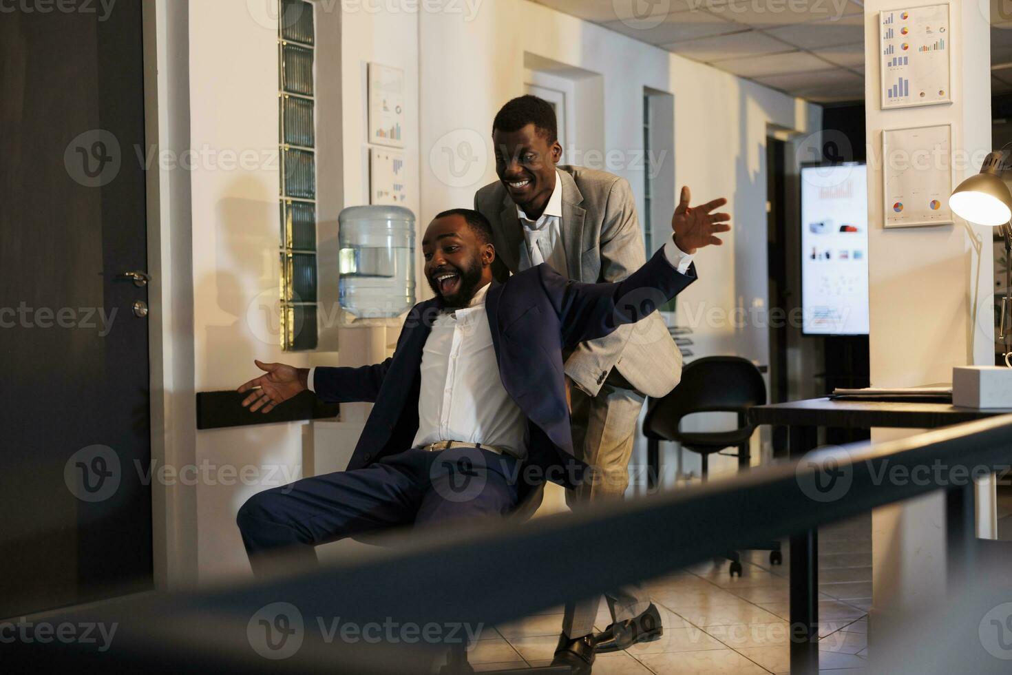 African american workers having sports race competition in modern startup office, taking break from work and having fun. Cheerful businessmen in formal suit riding desk chair late at night photo