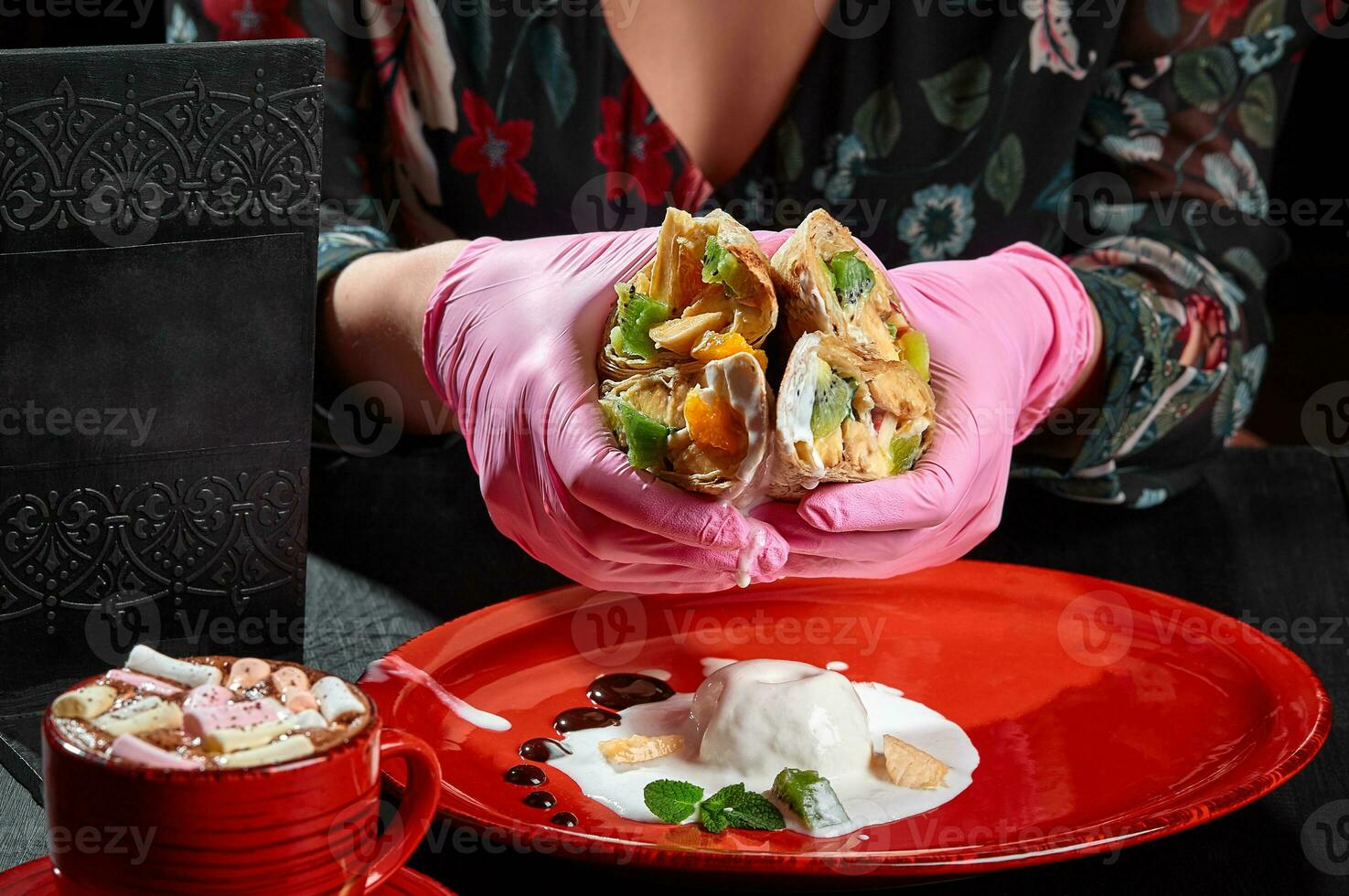 Female hands in rubber gloves holding fruit rolls in lavash over palte with ice cream and cup of cocoa photo