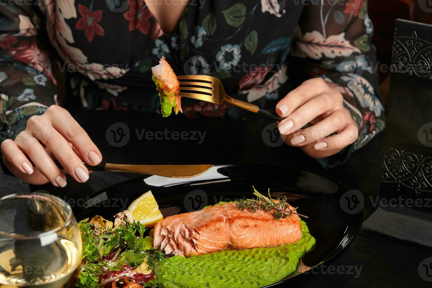 Female hands holding knife and fork with baked trout steak piece served with mashed peas, salad, lemon photo