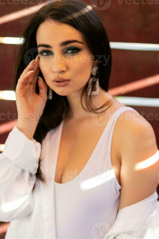 Glamorous young woman in white shirt posing near a wall photo