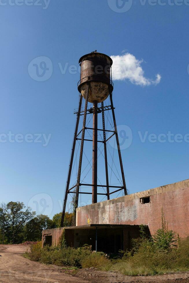 A beautiful water tower is set around an abandoned area. This rusty metal structure stands tall against a blue sky. photo