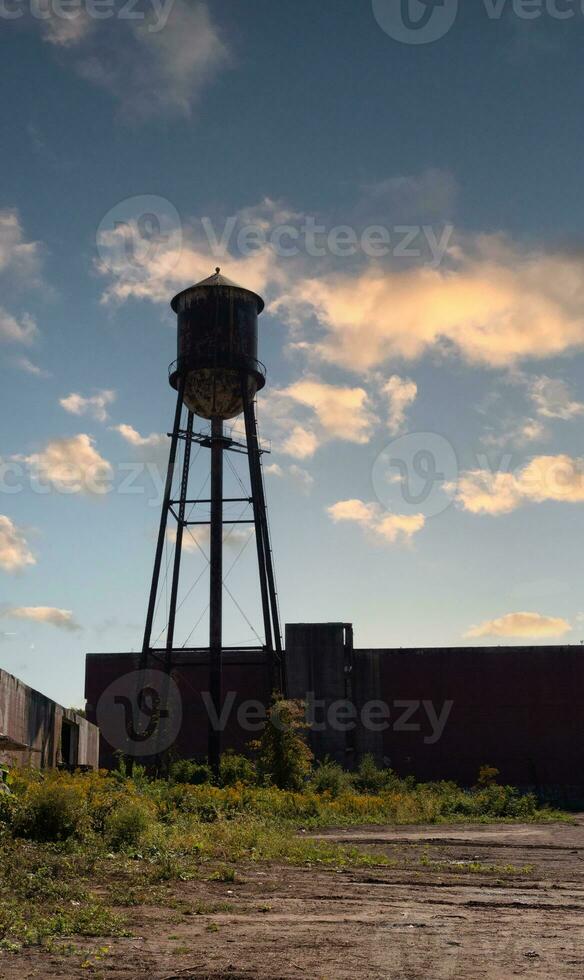 A beautiful water tower is set around an abandoned area. This rusty metal structure stands tall against a blue sky. photo