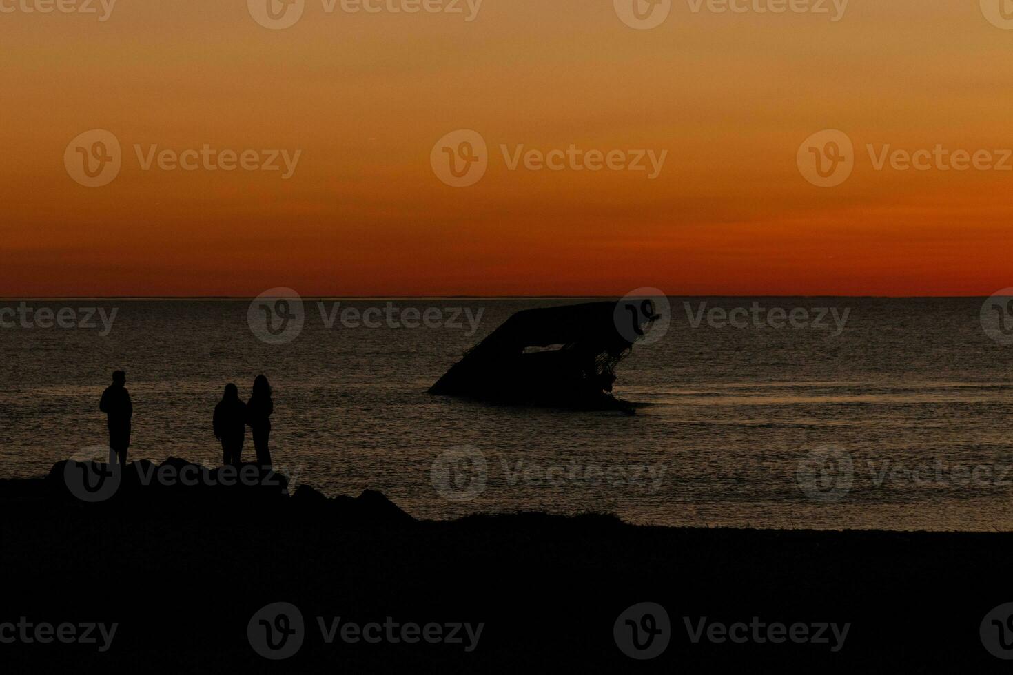 Sun setting on the Delaware Bay. The pretty red colors like the sky is on fire. The sunken ship of Cape May sitting in the ocean. People standing on the large black rocks of the jetty of the beach. photo