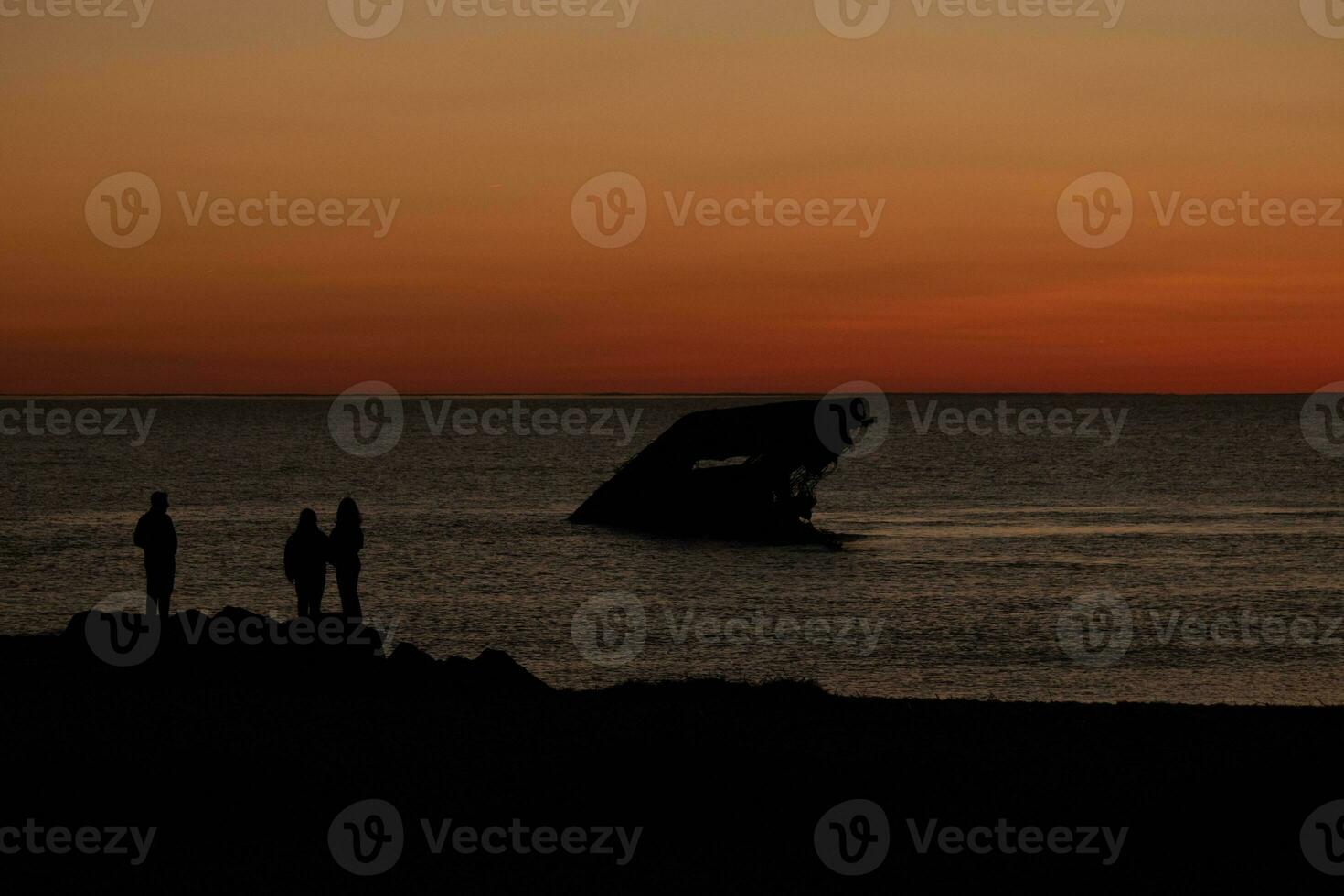 Sun setting on the Delaware Bay. The pretty red colors like the sky is on fire. The sunken ship of Cape May sitting in the ocean. People standing on the large black rocks of the jetty of the beach. photo