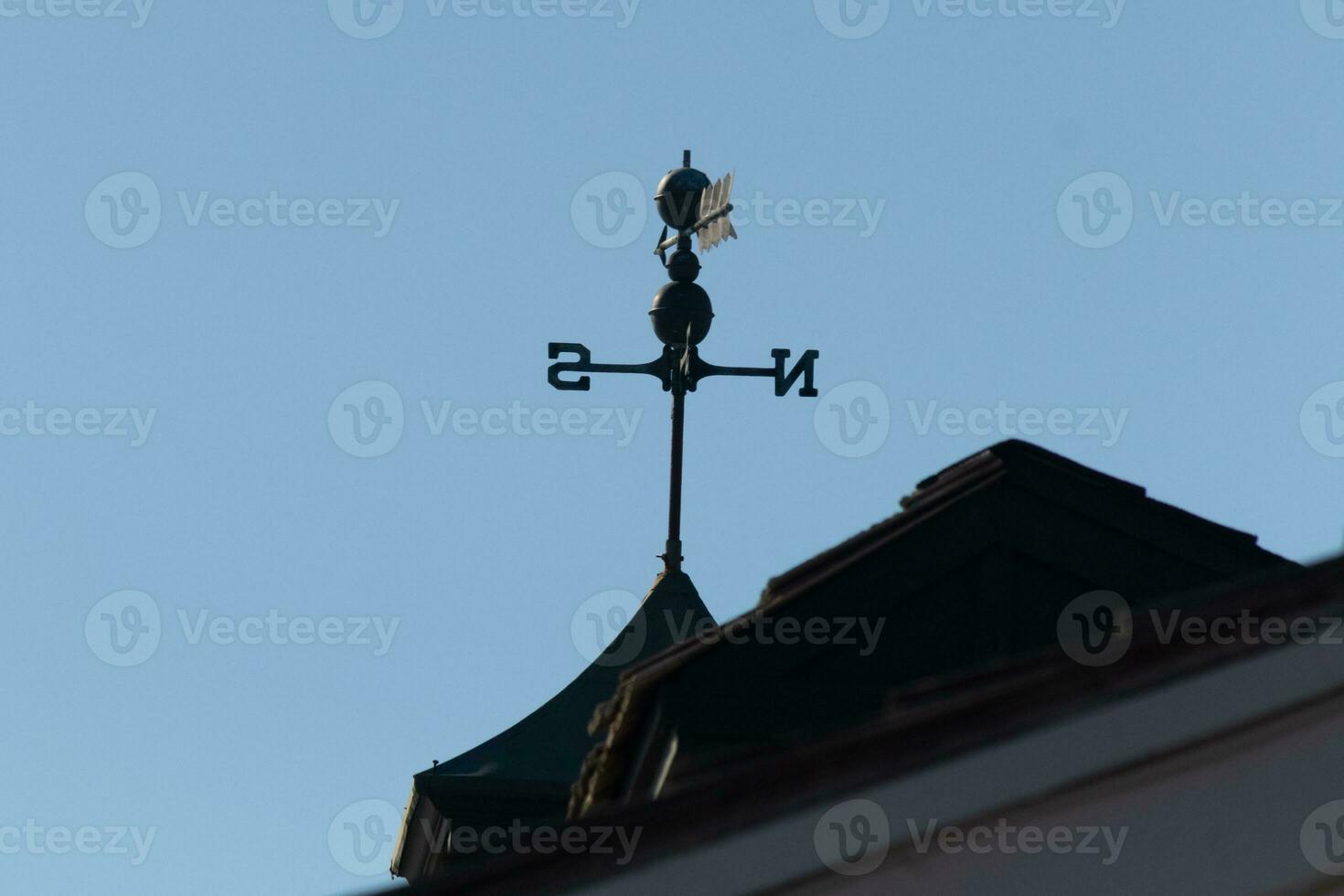 This weather vane was beautiful on the roof contrasted against the sky. The shadowy image with the clear blue sky in the background. The North, South, East, and West showing direction of the wind. photo