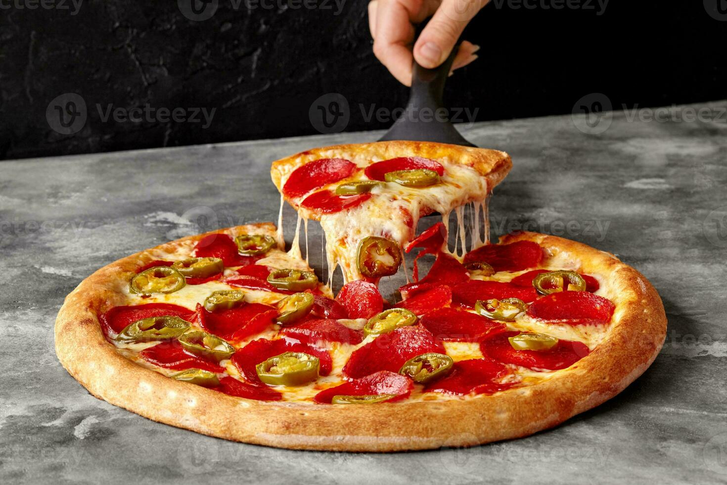 Female hand holding serving paddle with slice of pepperoni pizza with melted cheese and jalapeno photo