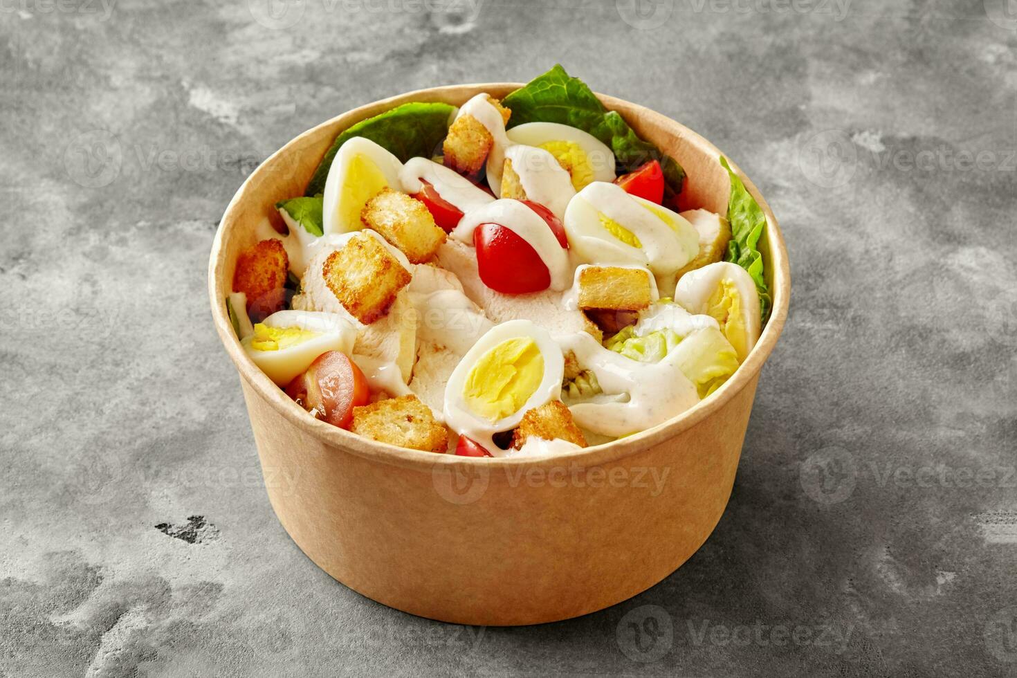 Paper bowl of Caesar salad with romaine lettuce, tomatoes, quail egg, croutons, chicken fillet and sauce photo