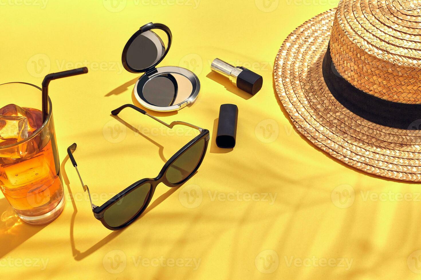Straw beach woman's hat, beverage with ice in a glass and sun glasses on yellow background with shadow from a palm leaf. photo