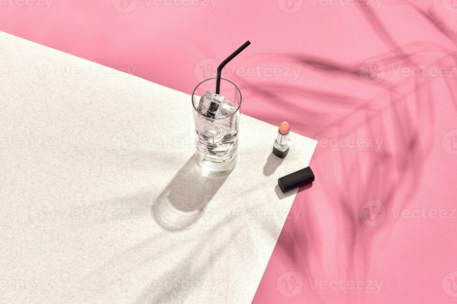 High angle view of lipstick and drinking glass with ice on table. Pink and white background with shadow from a palm leaf photo