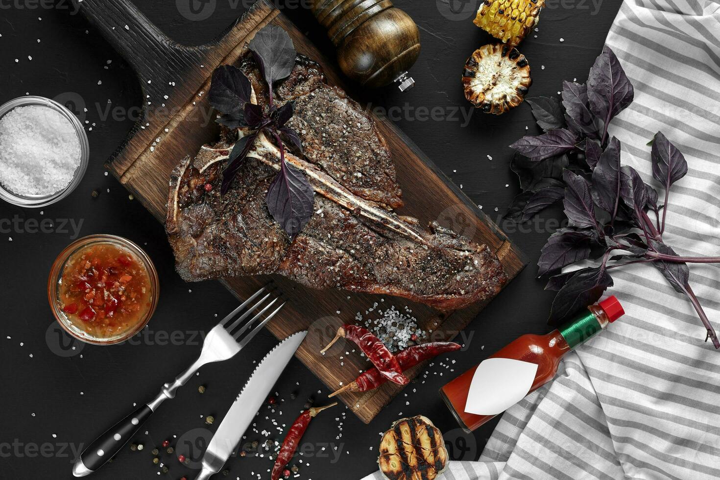 Succulent grilled large t-bone steak garnished with herbs, corn and salt with fork and knife beside it on cutting board photo