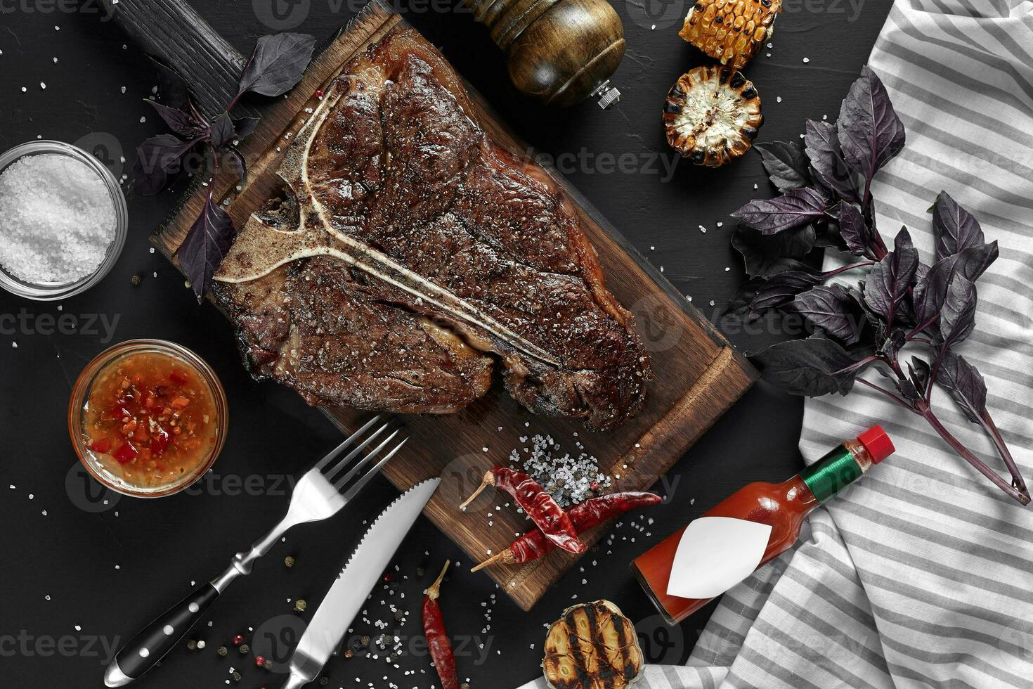Succulent grilled large t-bone steak garnished with herbs, corn and salt with fork and knife beside it on cutting board photo