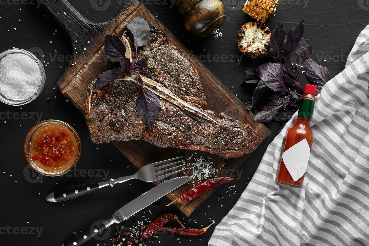 Succulent grilled large t-bone steak garnished with herbs, corn and salt with fork and knife beside it on cutting board photo