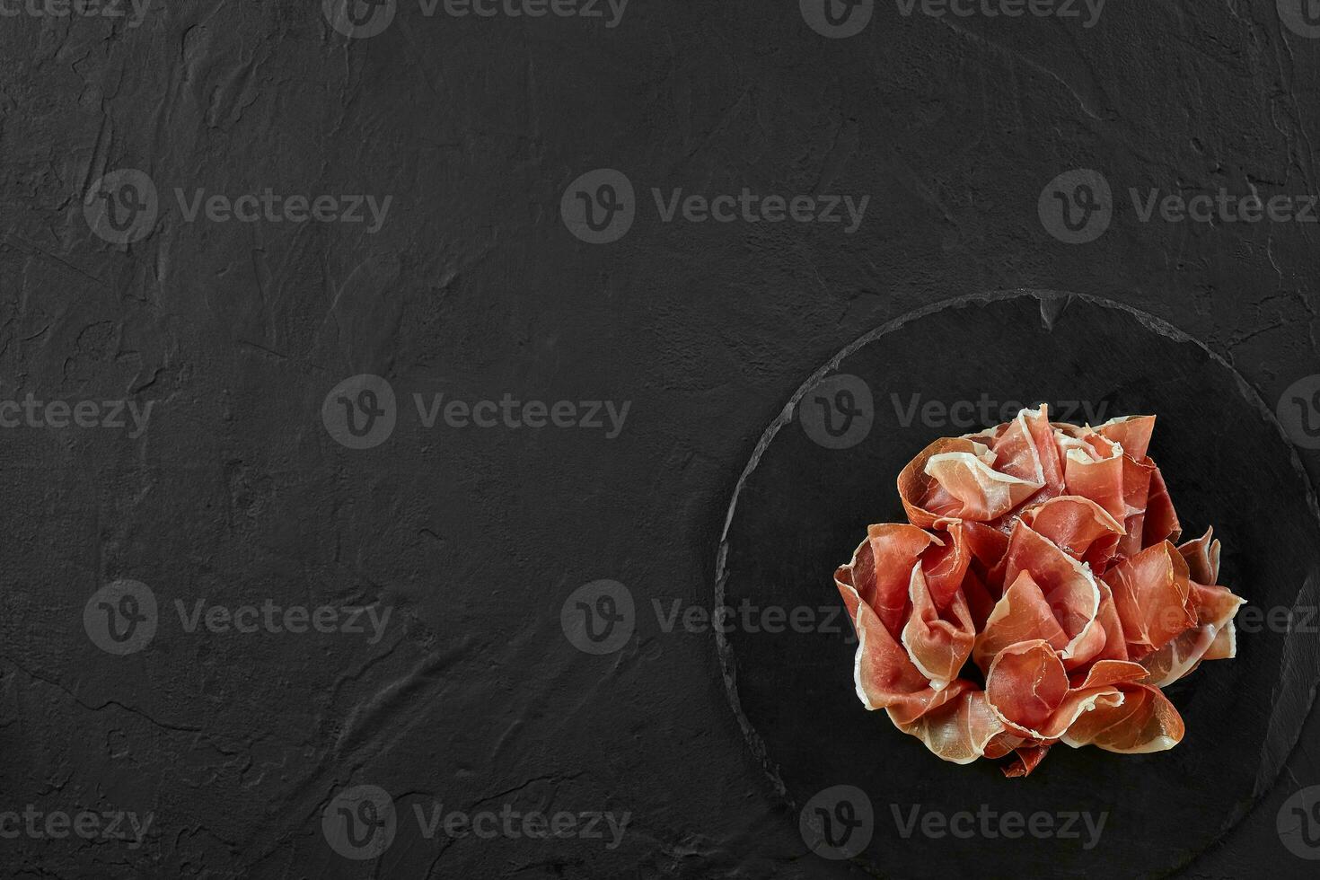 Gourmet, manually sliced jamon on black stone slate board against a dark grey background. Close-up shot. Top view. Copy space. photo