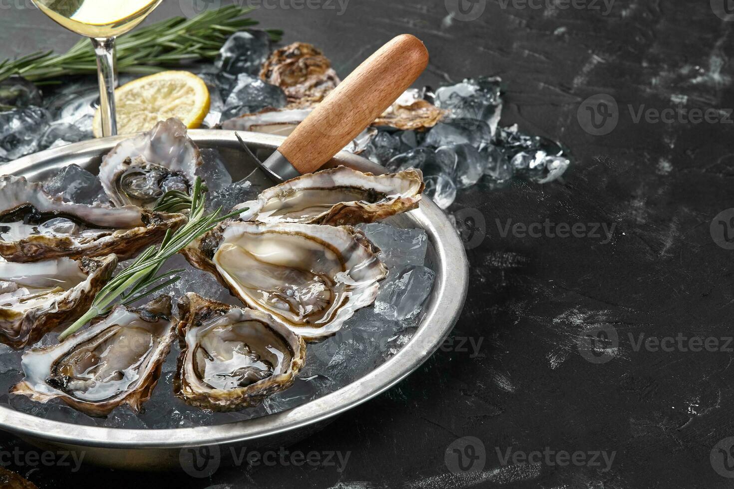 Fresh opened oysters, ice, lemon on a round metal plate and champagne are on a black stone textured background. Side view with copy space. Close-up. photo