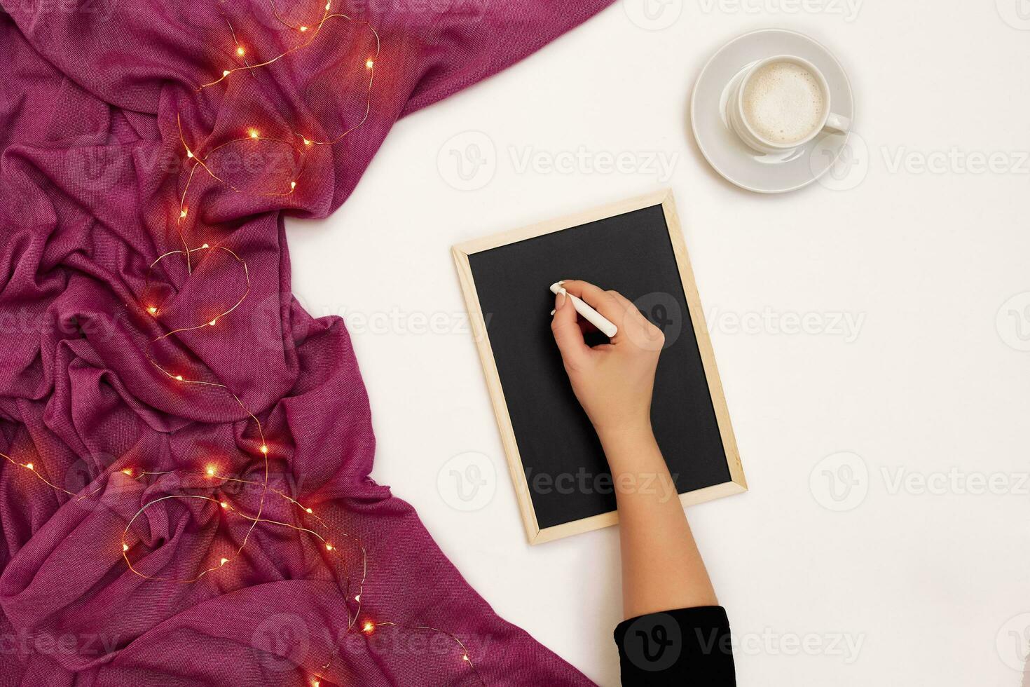 Top view of girl's hand writing on small black wooden board on white surface with coffee cup and other items. Mock up photo