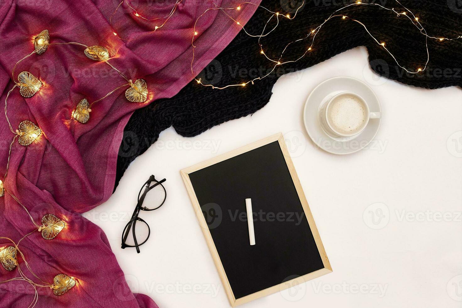 A cup of coffee, a small black wooden board and a white chalk with new years goals. Scarf and Christmas lights on white background. photo